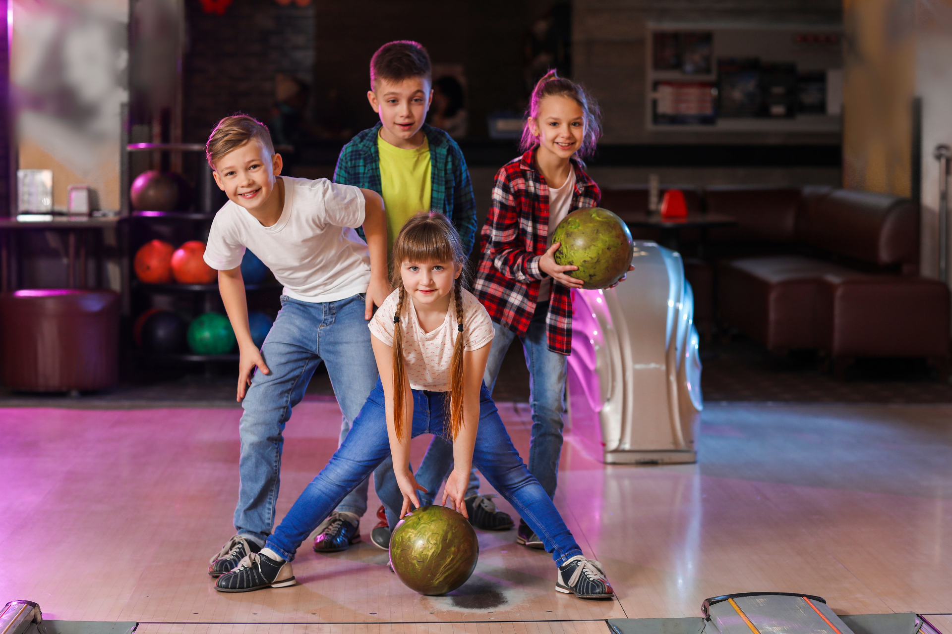 Four children in a bowling alley, smiling and posing with bowling balls on the lane