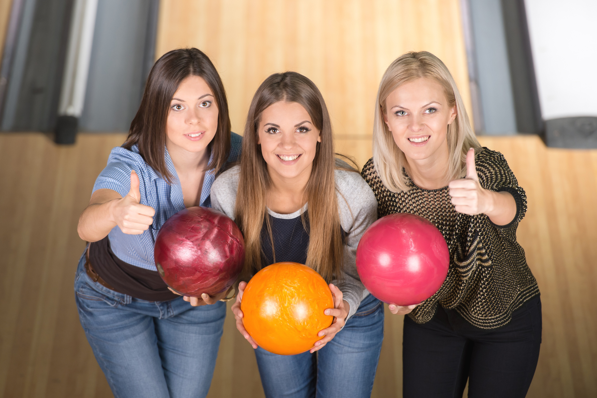 Three people holding colorful bowling balls, smiling in a bowling alley