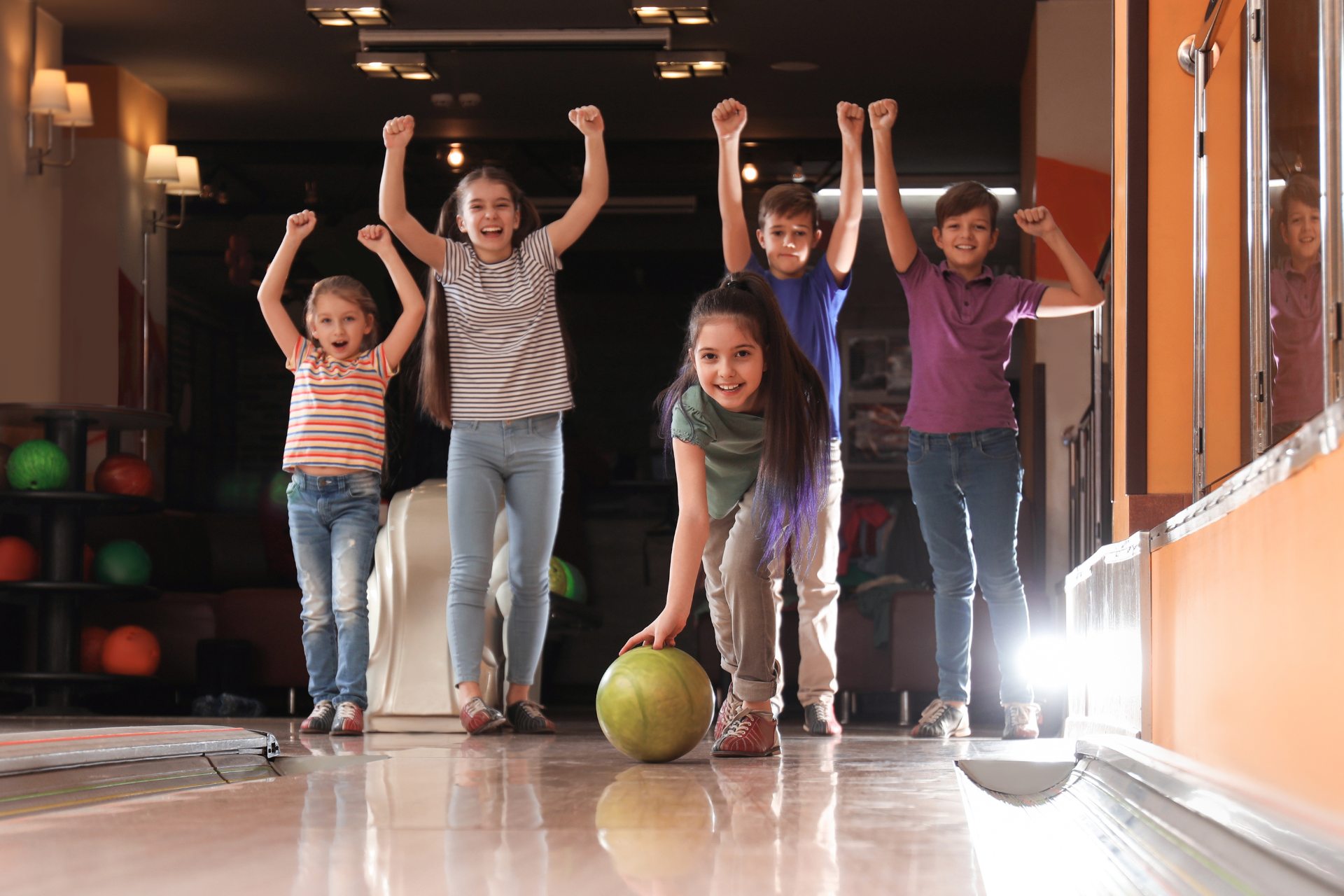 Four children cheer beside a bowling lane, with one child rolling a green ball toward the pins.
