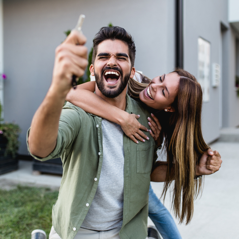 Excited couple taking a selfie with a key in his hand from buying a new home, with the woman hugging the man from behind.