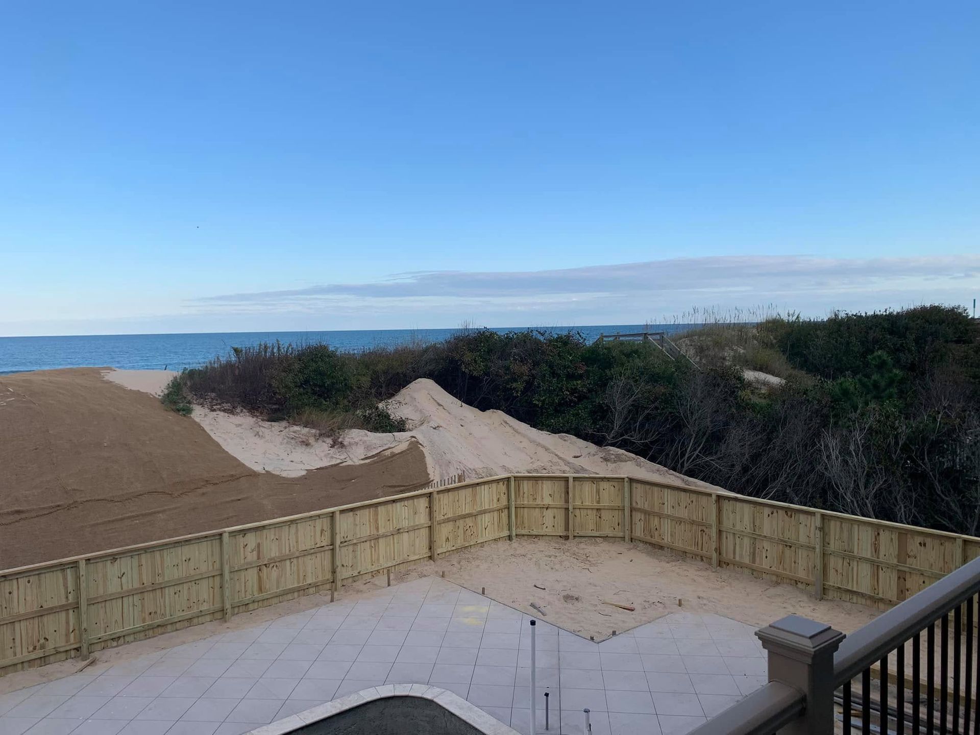 Sandy coastal yard with wooden fence, deck, and ocean view under a clear blue sky