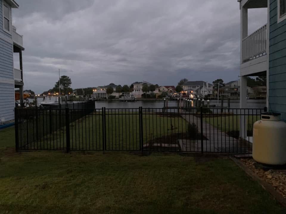 Cloudy waterfront view through a black fence, with grassy yard and apartment buildings on both sides. Cloudy waterfront view through a black fence, with grassy yard and apartment buildings on both sides.