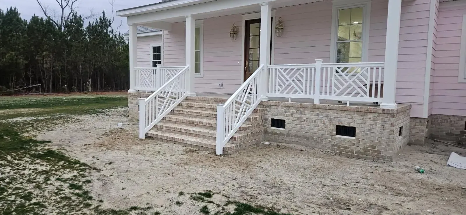 Pink house porch with white railings and steps on a dirt lot, viewed from the front