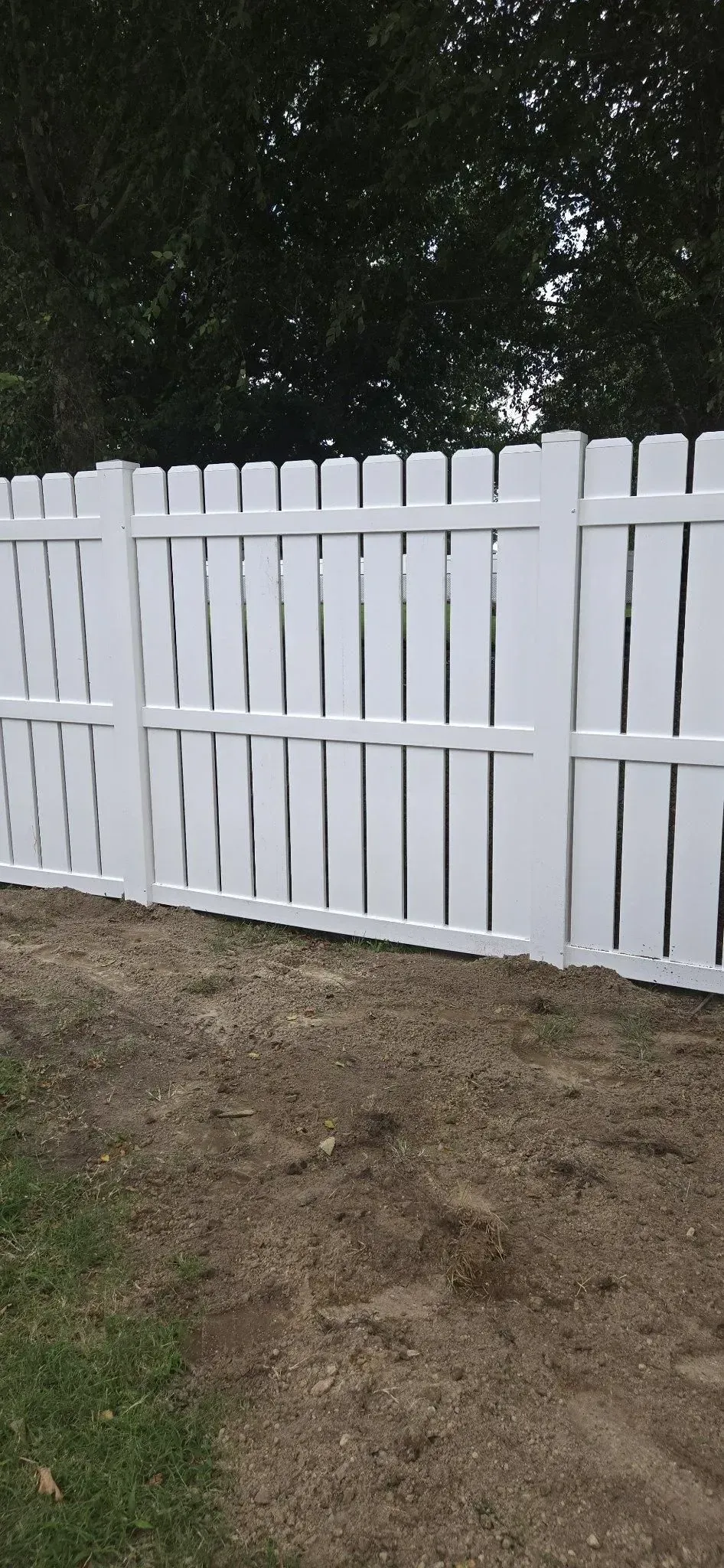 White wooden fence with a gate beside a dirt yard and trees in the background