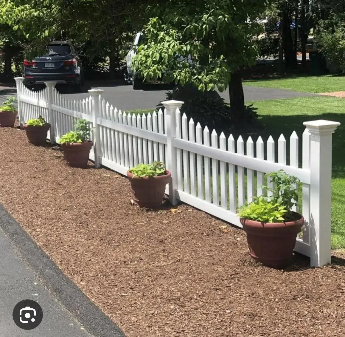 White picket fence with potted plants lining a mulched driveway beside a parked car and leafy yard