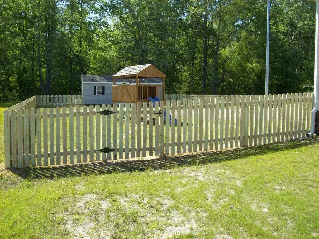 White picket fence around a small playhouse in a grassy yard with trees in the background