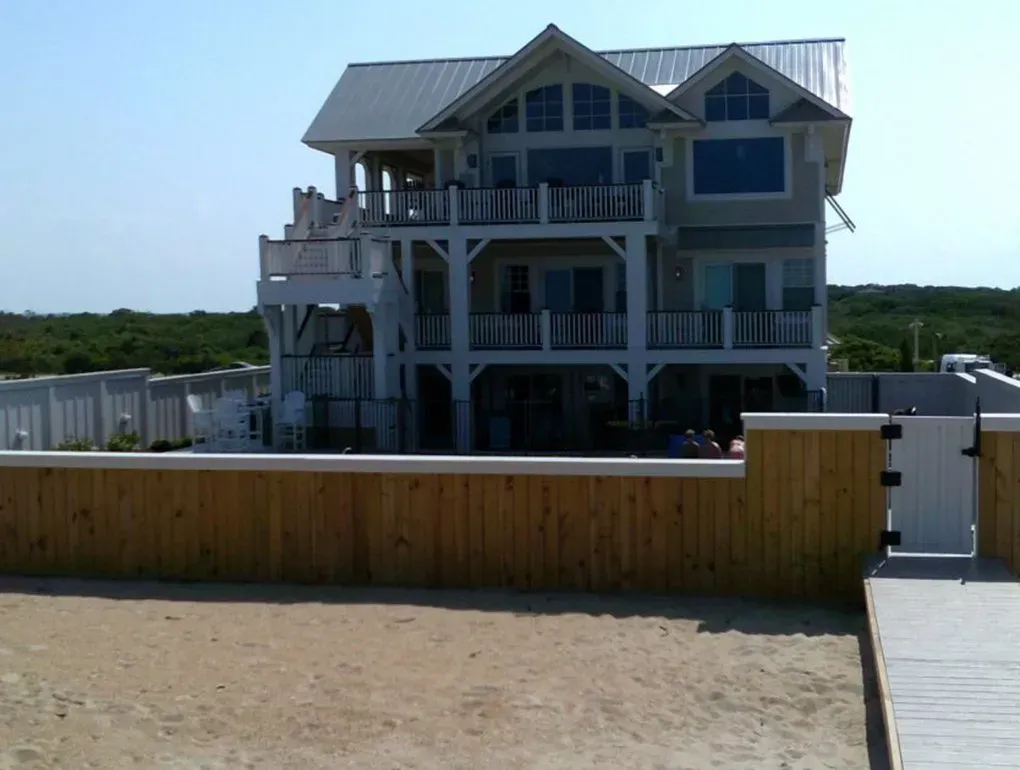 Beach house with white and blue exterior behind a wooden fence and sandy foreground.