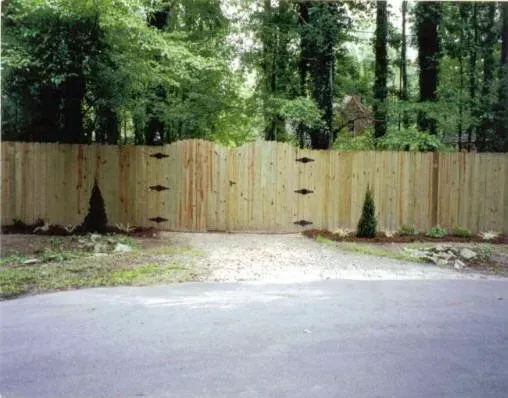 Wooden gate in a fenced driveway entrance, backed by dense green trees.