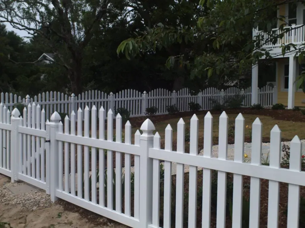 White picket fence along a garden path in front of a house, with trees and a porch in the background