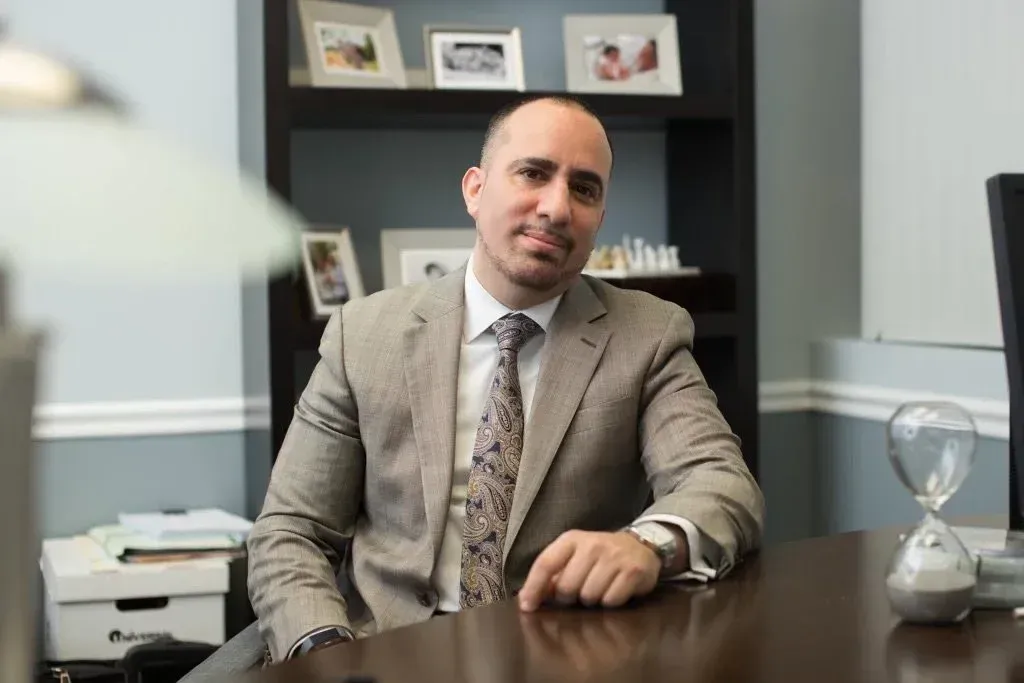 Man in a suit sits at a desk in an office with shelves and family photos.