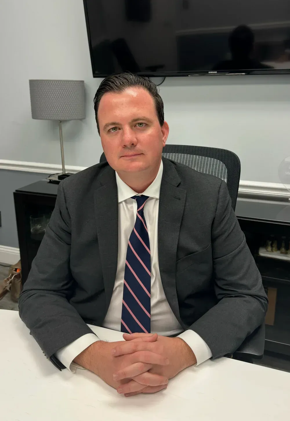 Man in a suit, seated at a desk, arms crossed. He is looking at the camera. Neutral expression.
