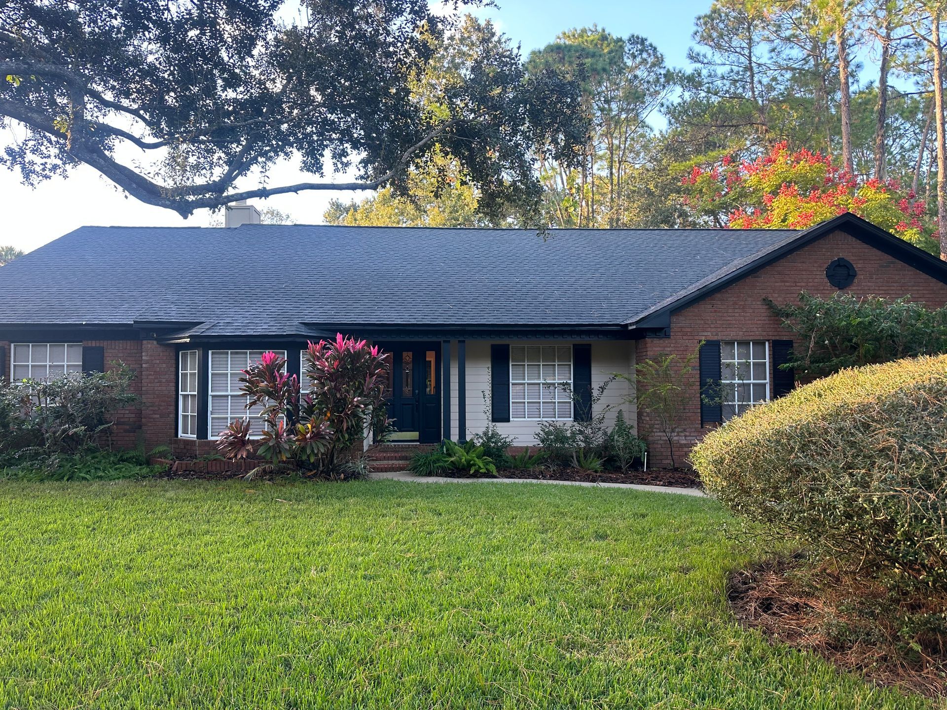 Ranch-style house with dark roof and brick facade, surrounded by green lawn and trees.