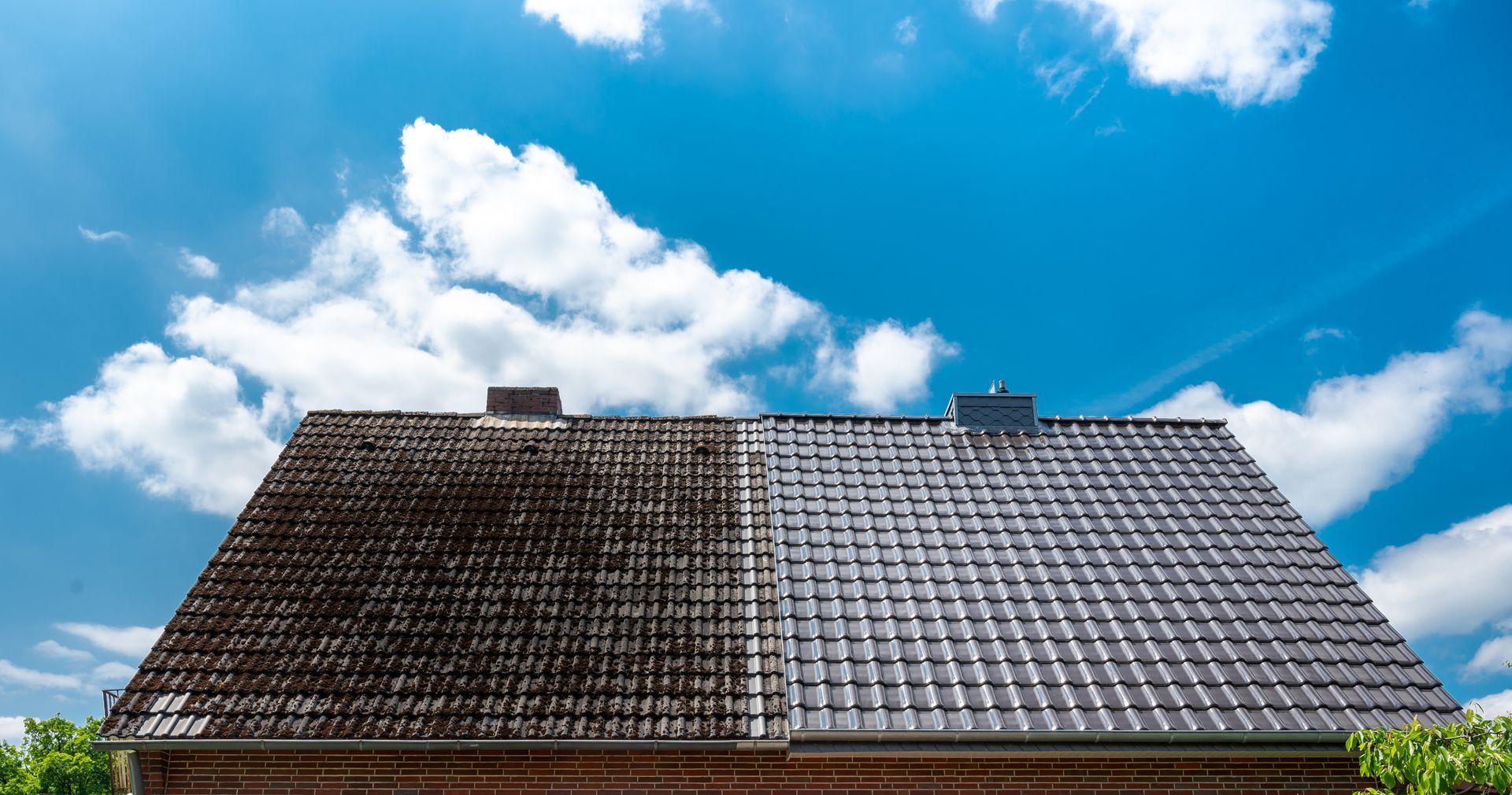 Roof of a house, half dirty and half clean, against a bright blue sky with puffy white clouds.
