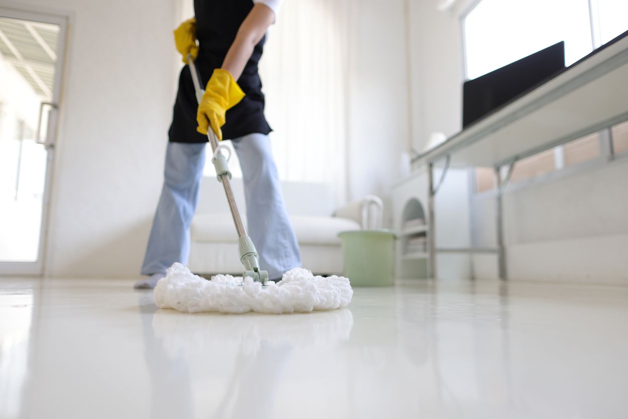Person mopping a shiny, white floor in a brightly lit room. They wear yellow gloves and an apron.