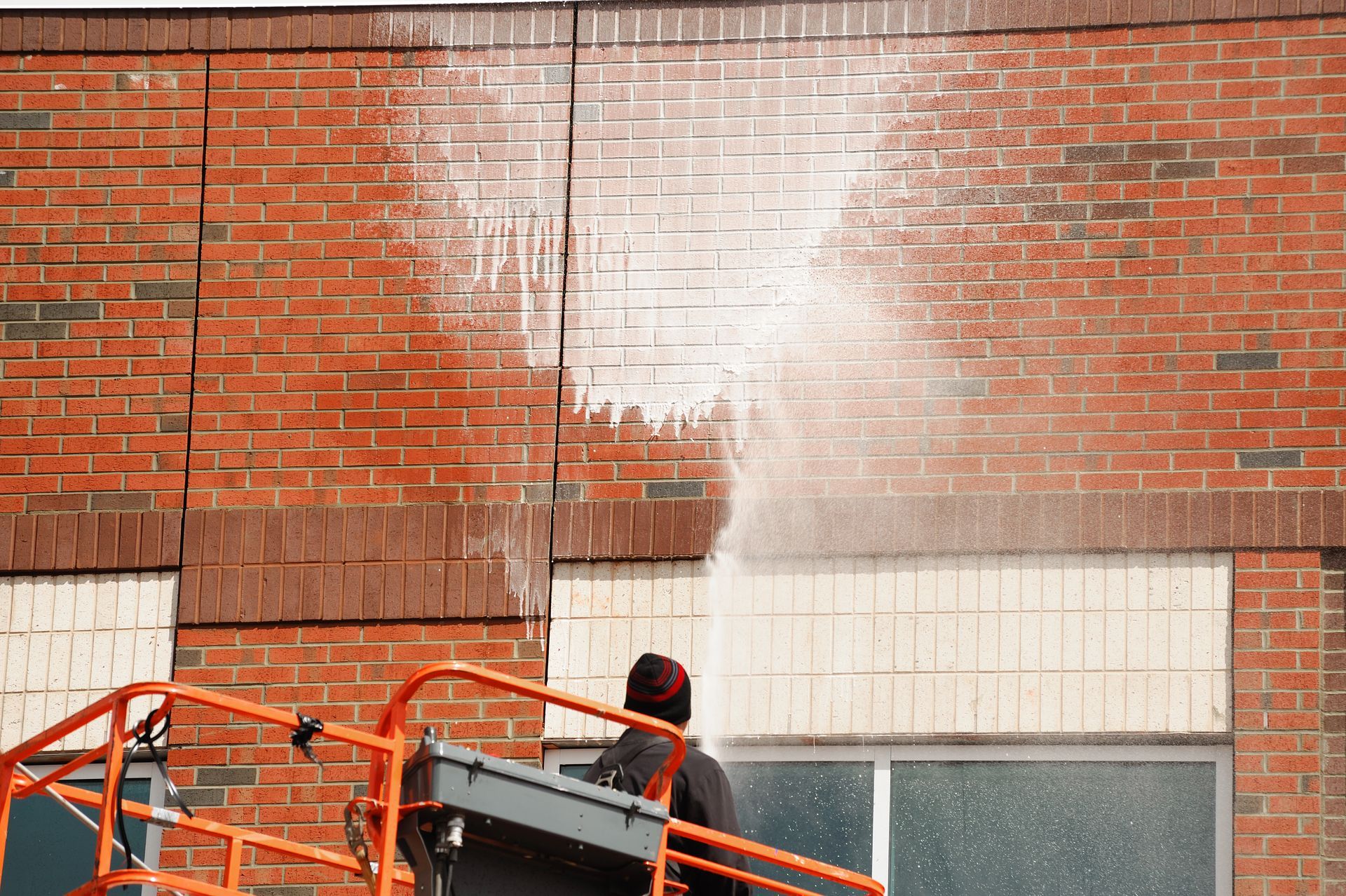 Man on a lift sprays water onto a brick building exterior, cleaning the wall.