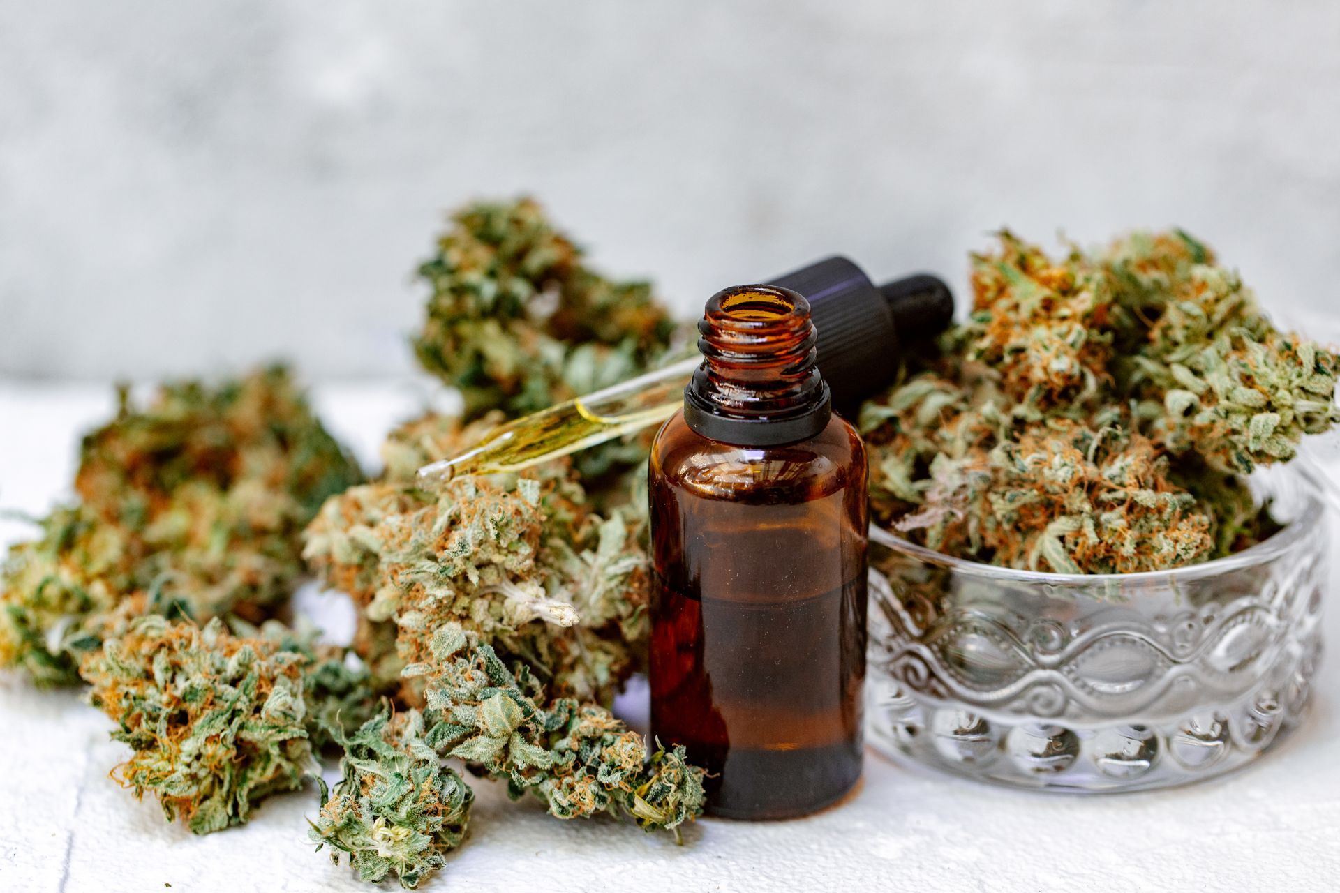 Amber glass dropper bottle beside cannabis buds in a silver bowl on a white surface