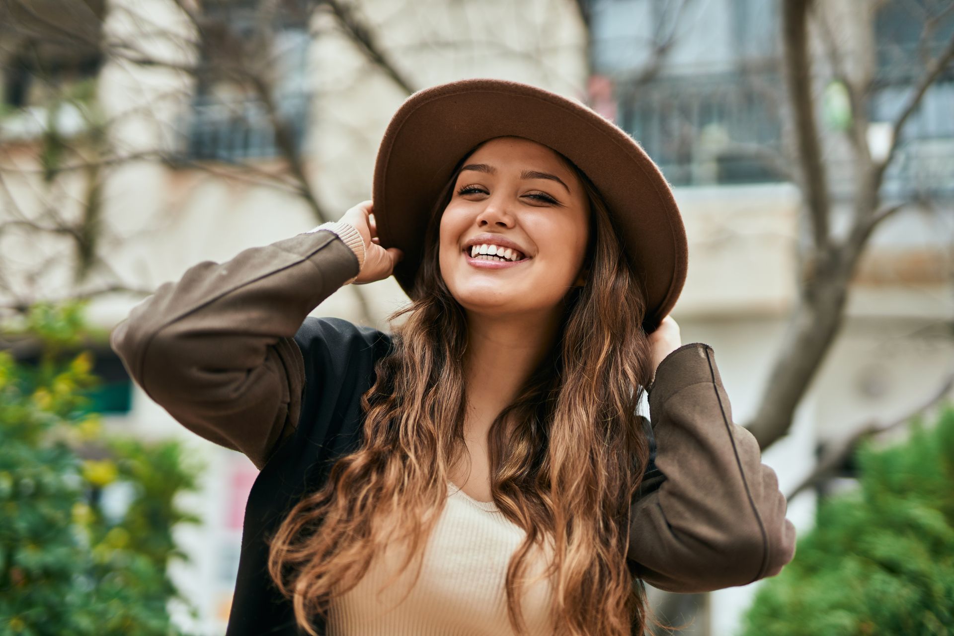 Smiling person in a brown hat and jacket outdoors, adjusting the hat with both hands.