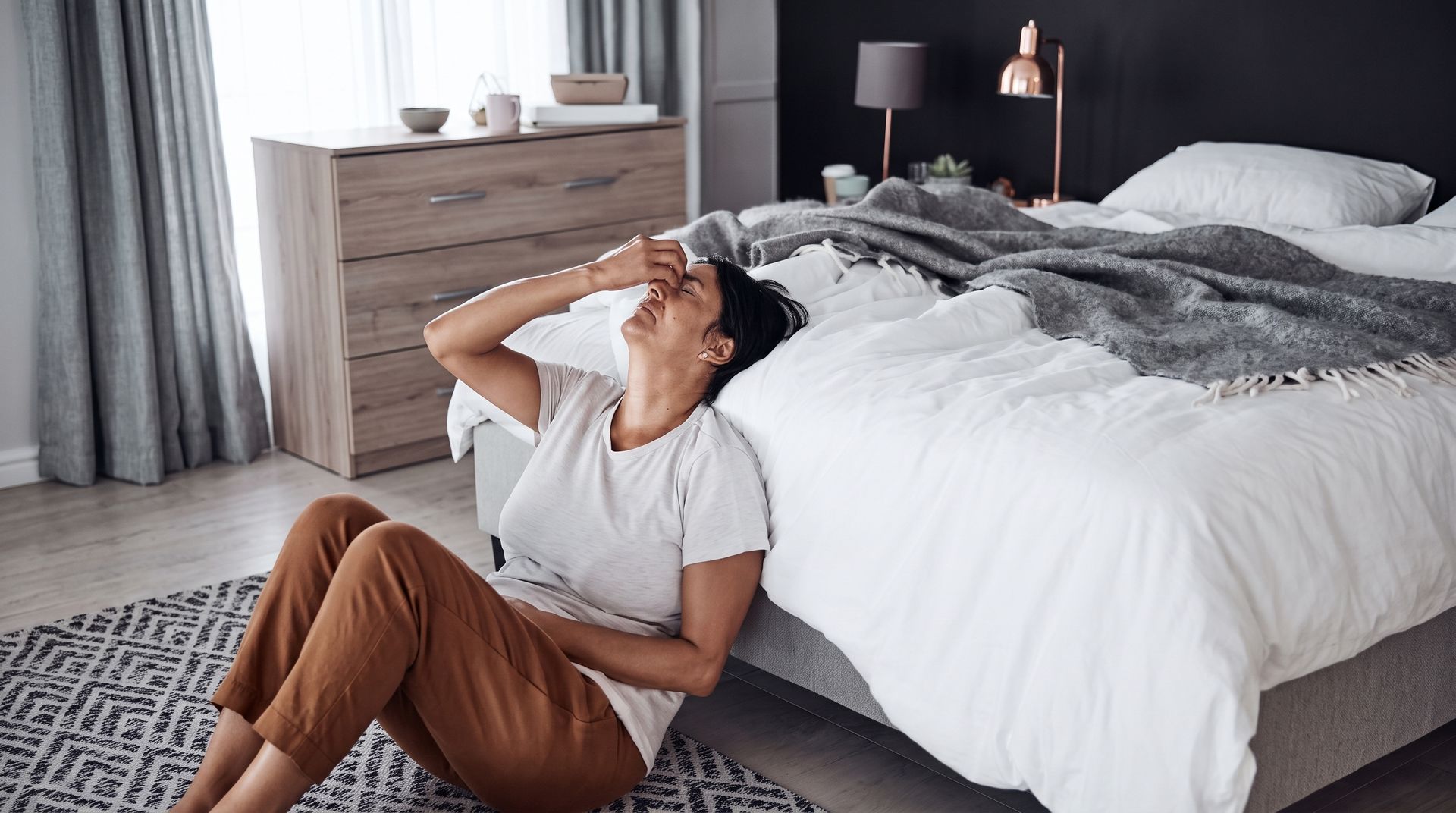 Woman sitting curled up on a bed, hugging her knees and looking down sadly in a softly lit room