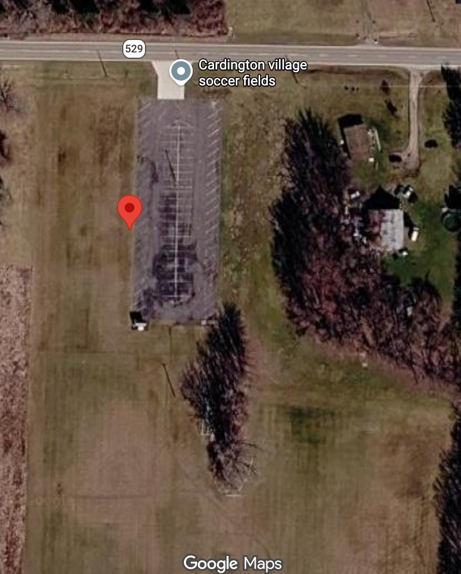 Google Maps satellite view of a rural building beside a road, with a red location pin on open grass.