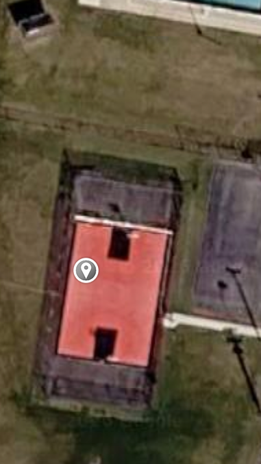 Aerial view of a red-roofed building with a dark roof section, beside a grassy area and pavement.