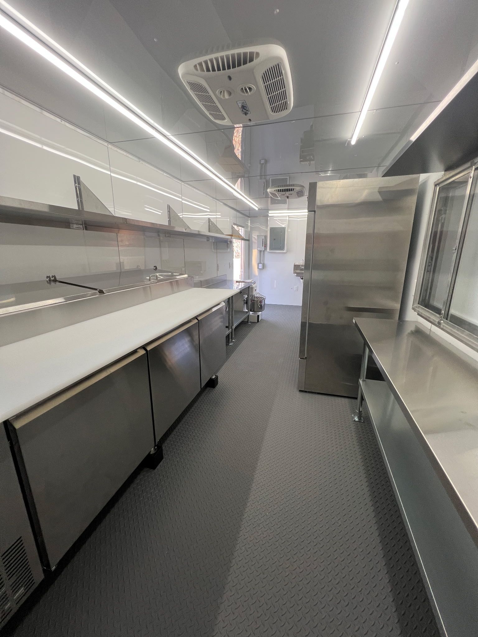 Narrow stainless-steel galley aisle with shelves, cabinets, and overhead lights in a modern cargo trailer interior
