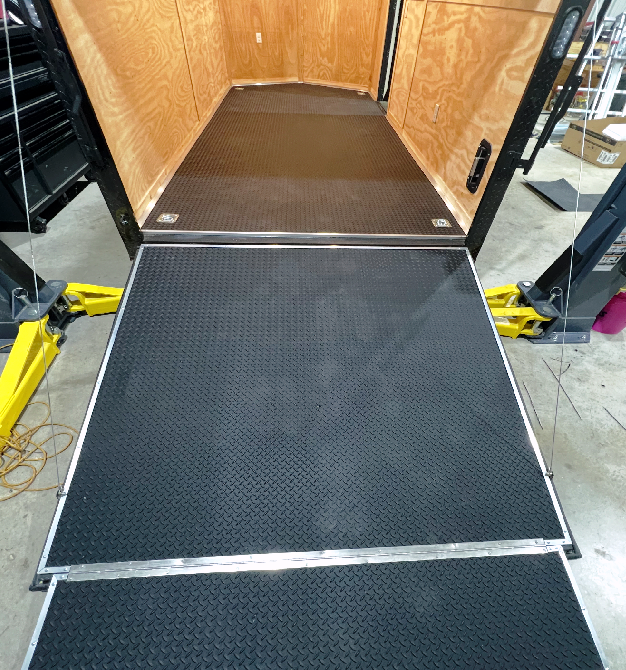 Black ramp leading into a trailer with tan wooden walls and yellow lift supports on each side.