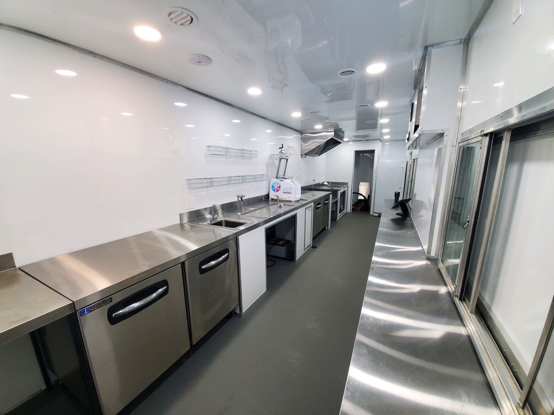 Bright commercial kitchen corridor with stainless counters, sink, ovens, and white walls under recessed lights