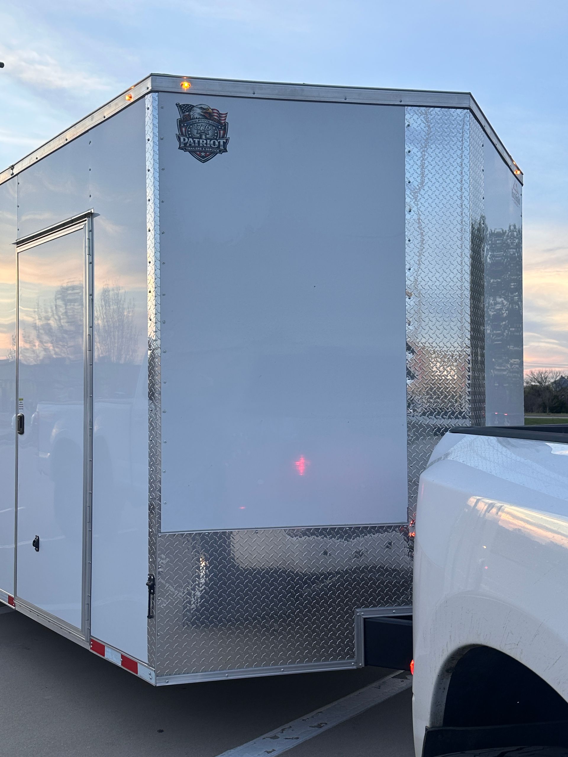 White enclosed trailer with silver diamond-plate trim parked beside a white pickup truck at sunset