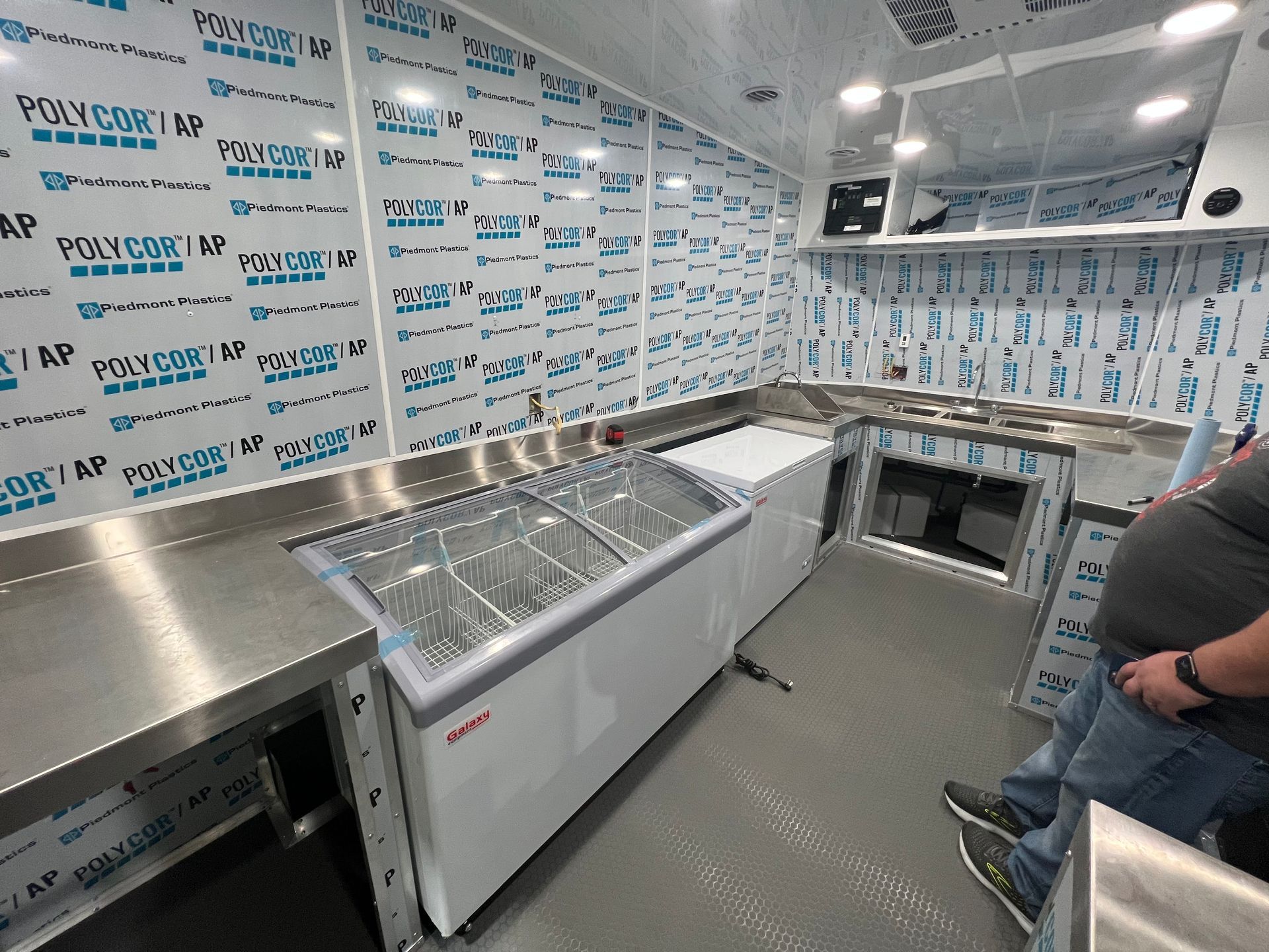 Stainless-steel prep area with freezers and a person seated on the right in a clean, white-walled kitchen trailer.