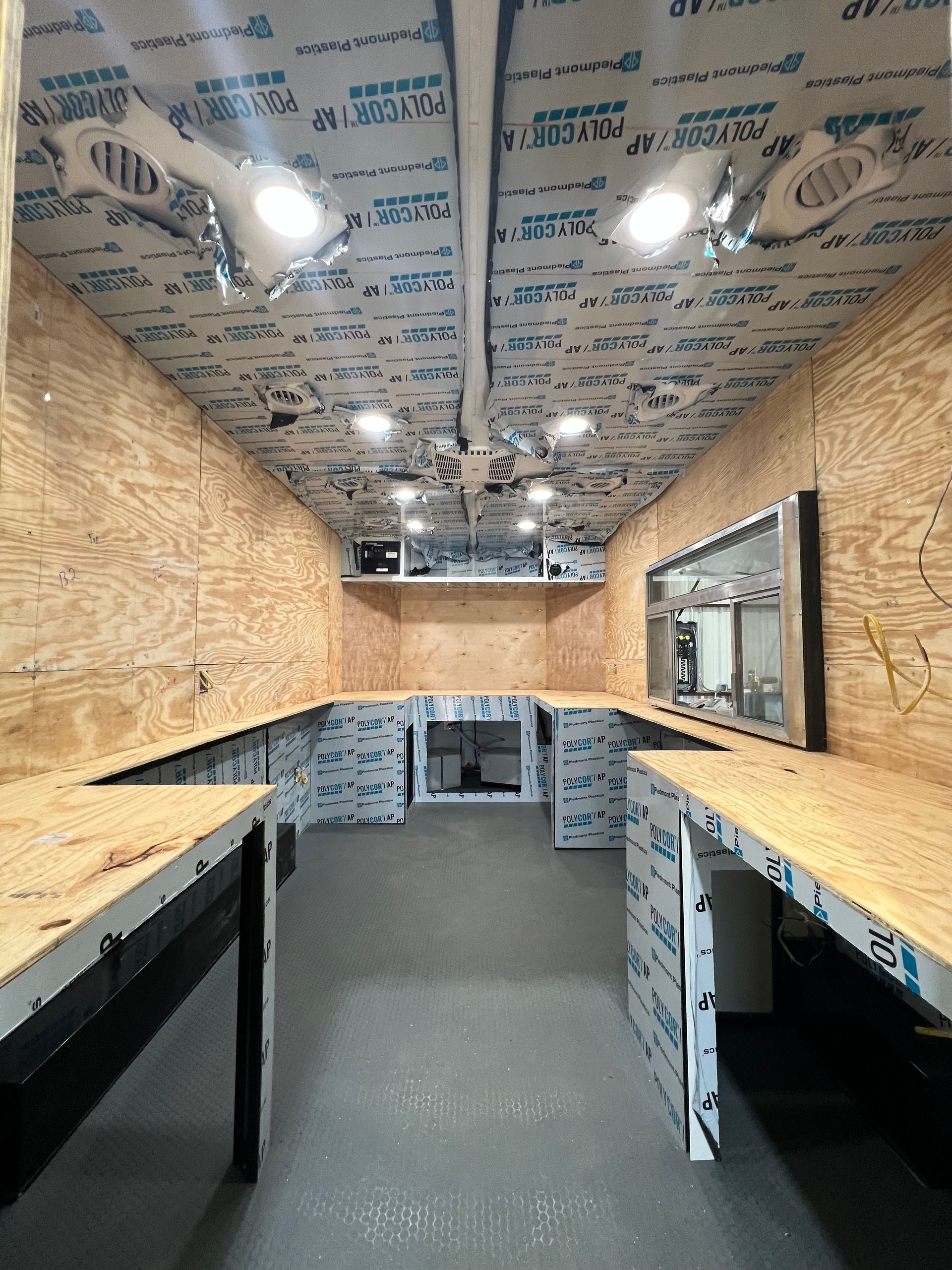 Narrow workshop van interior with plywood walls, gray floor, and workbenches along both sides.