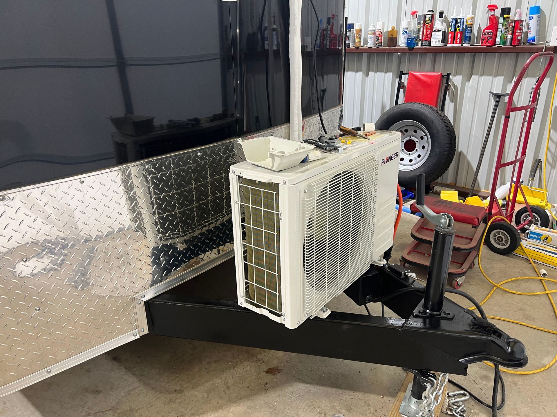 White HVAC unit mounted on a black trailer hitch in a garage, with a spare tire and tools in the background