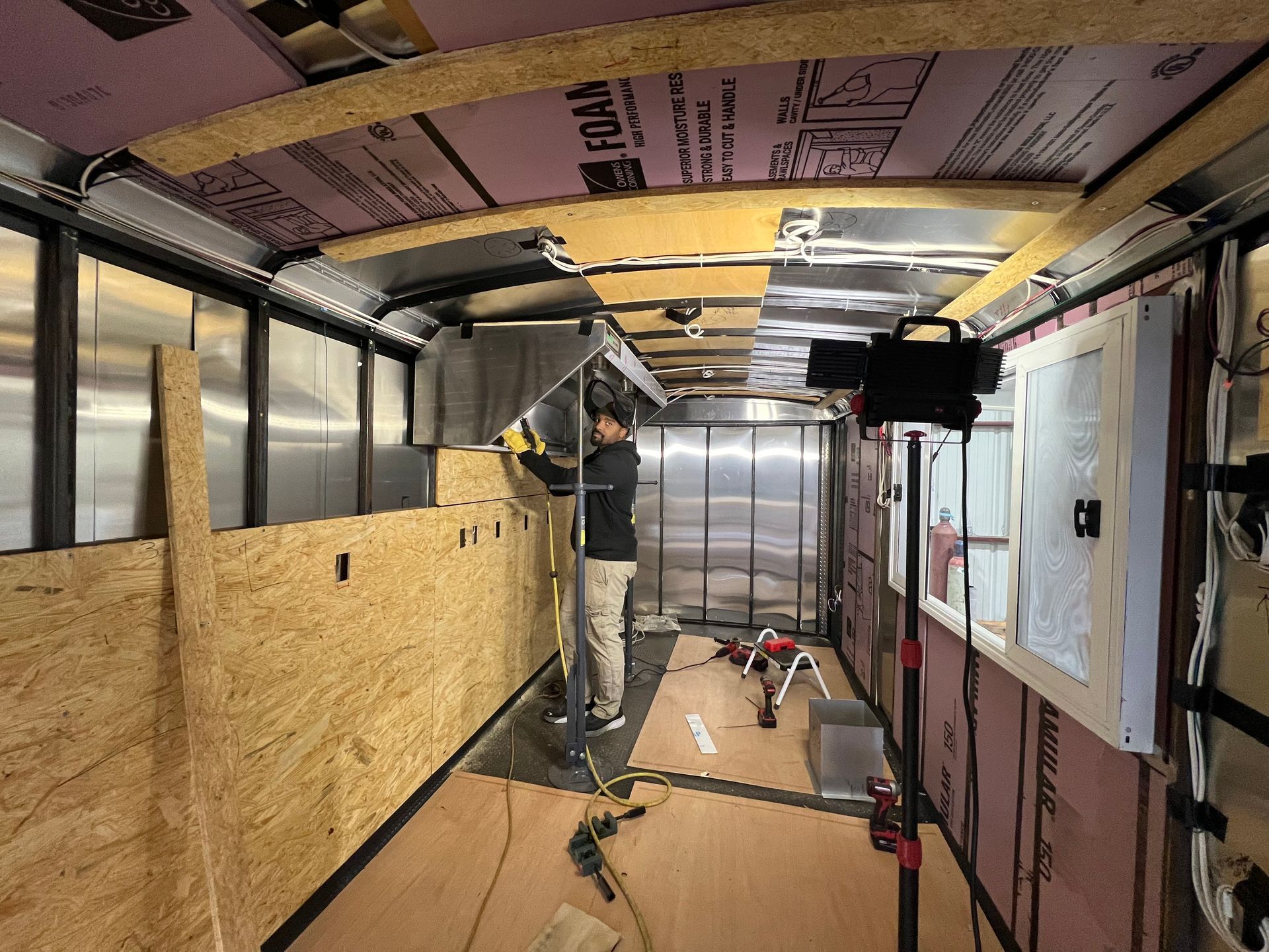 Worker installing insulation inside a partially upgraded trailer interior with exposed wood, metal framing, and wiring.