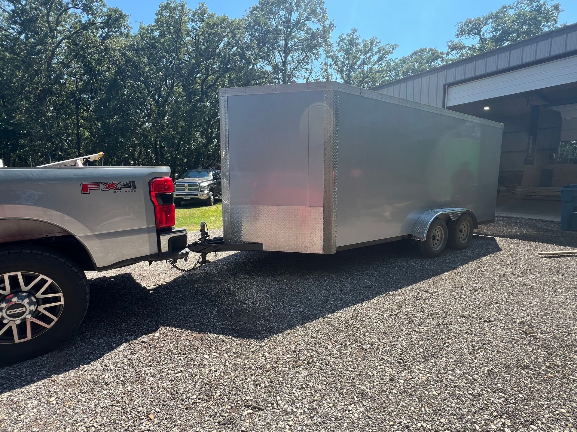 Silver enclosed cargo trailer hitched to a white pickup truck, parked on gravel beside a building.