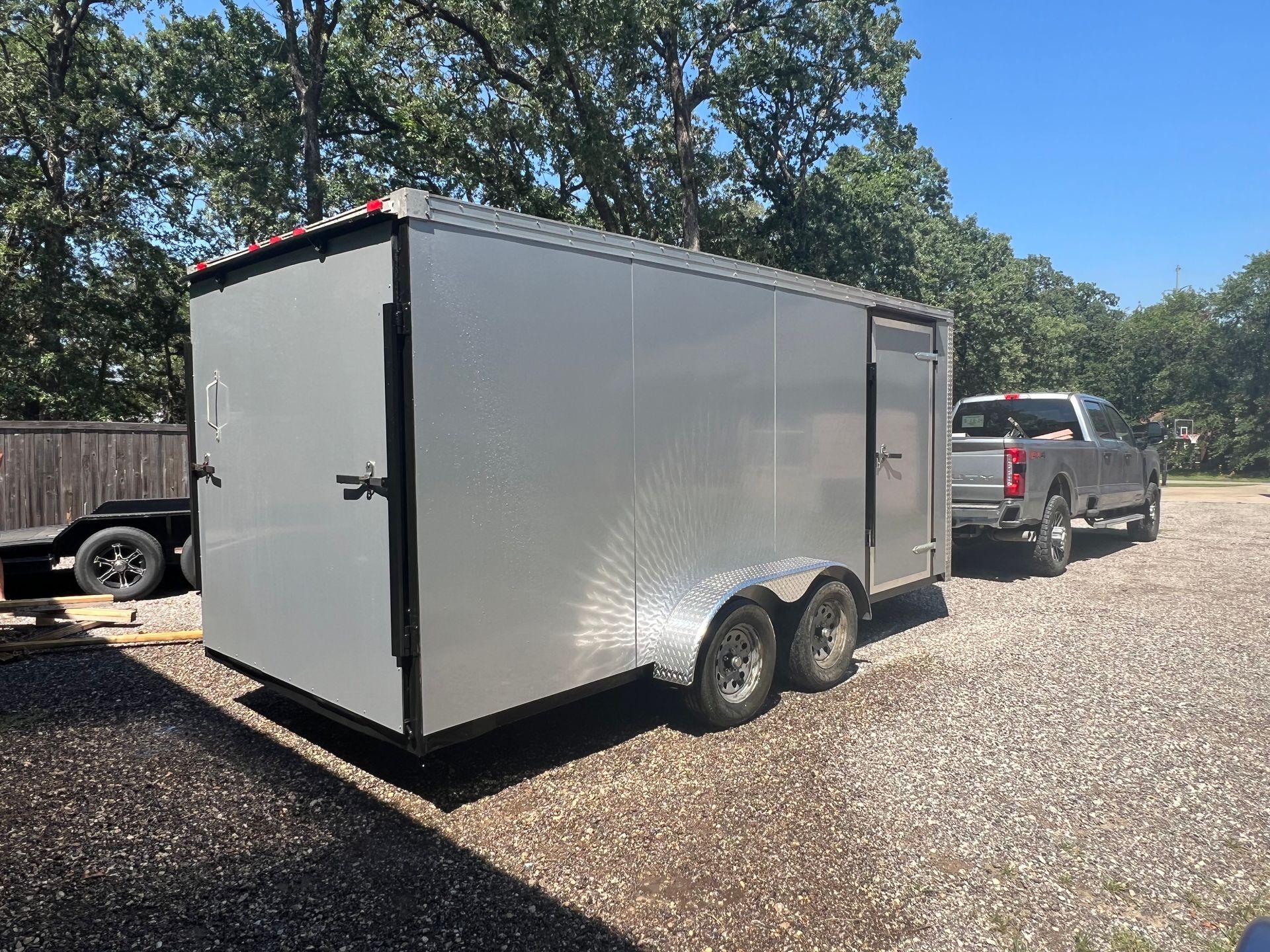 White enclosed cargo trailer hitched to a silver pickup truck on a gravel driveway under trees.