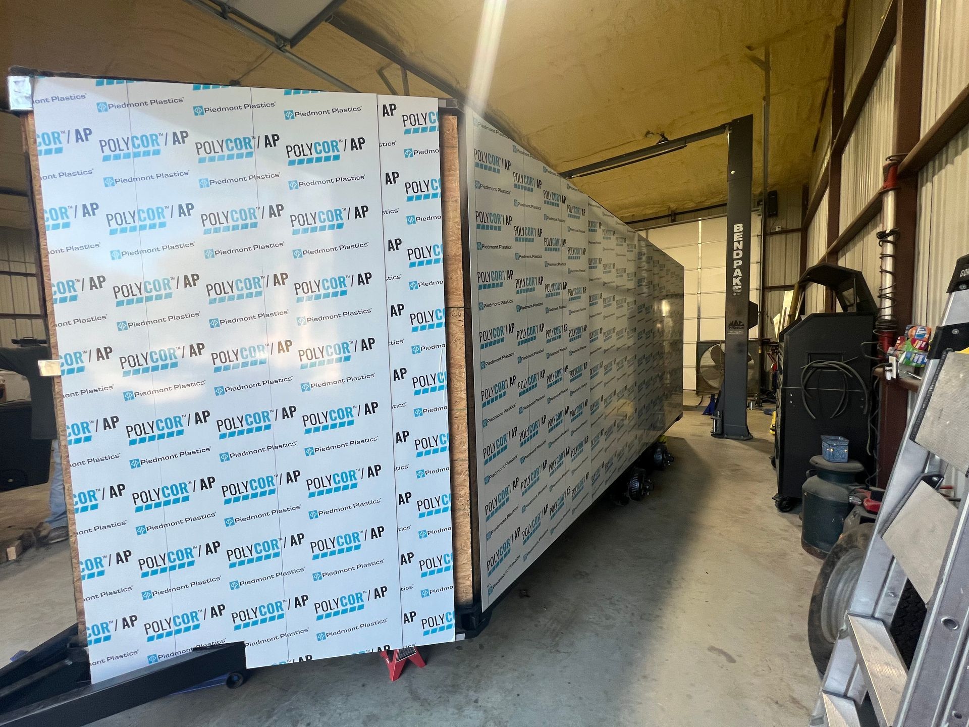 Large white-wrapped shipping crate on a warehouse floor, with workers and equipment in the background.