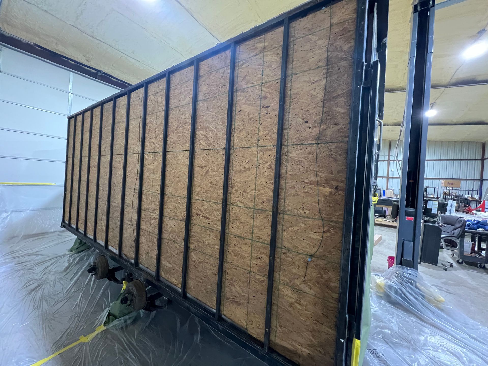 large brown cargo trailer in an industrial workshop, wrapped in plastic at one end.