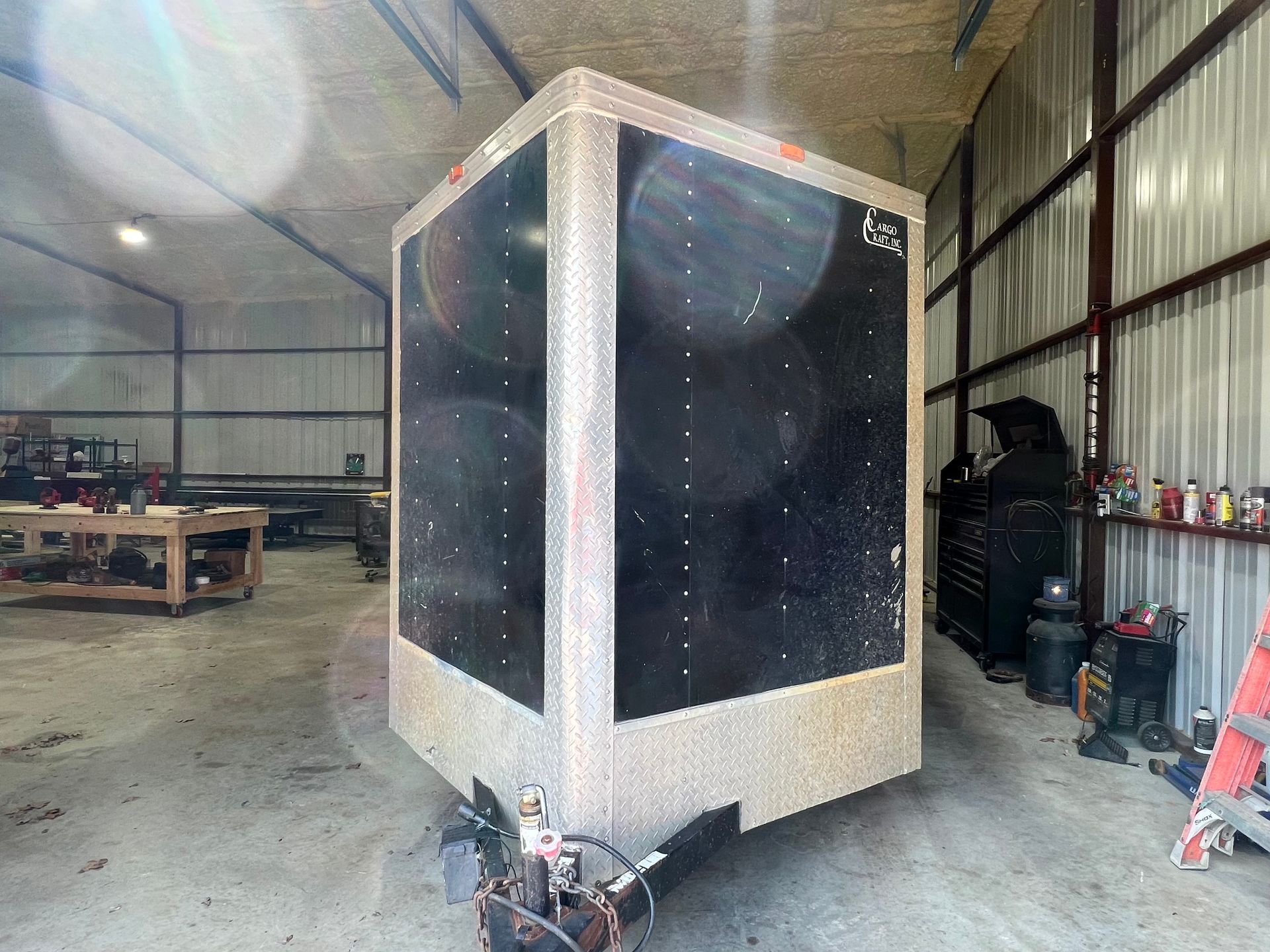 Large black-and-silver enclosed trailer in a warehouse, with lights and tools nearby.