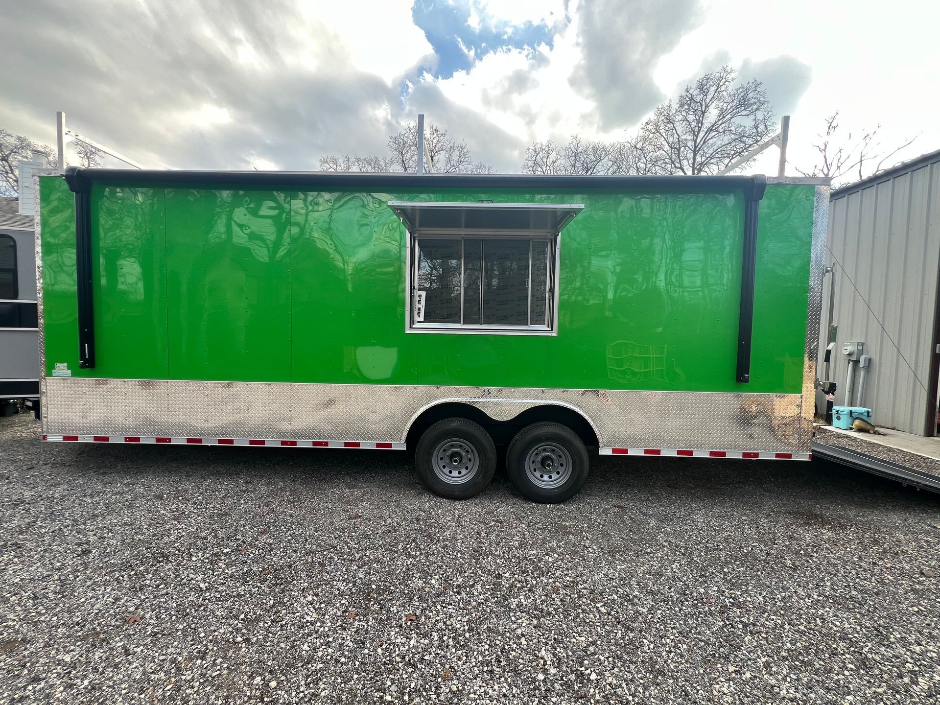 Bright green food trailer with a window, parked on gravel next to a white building.