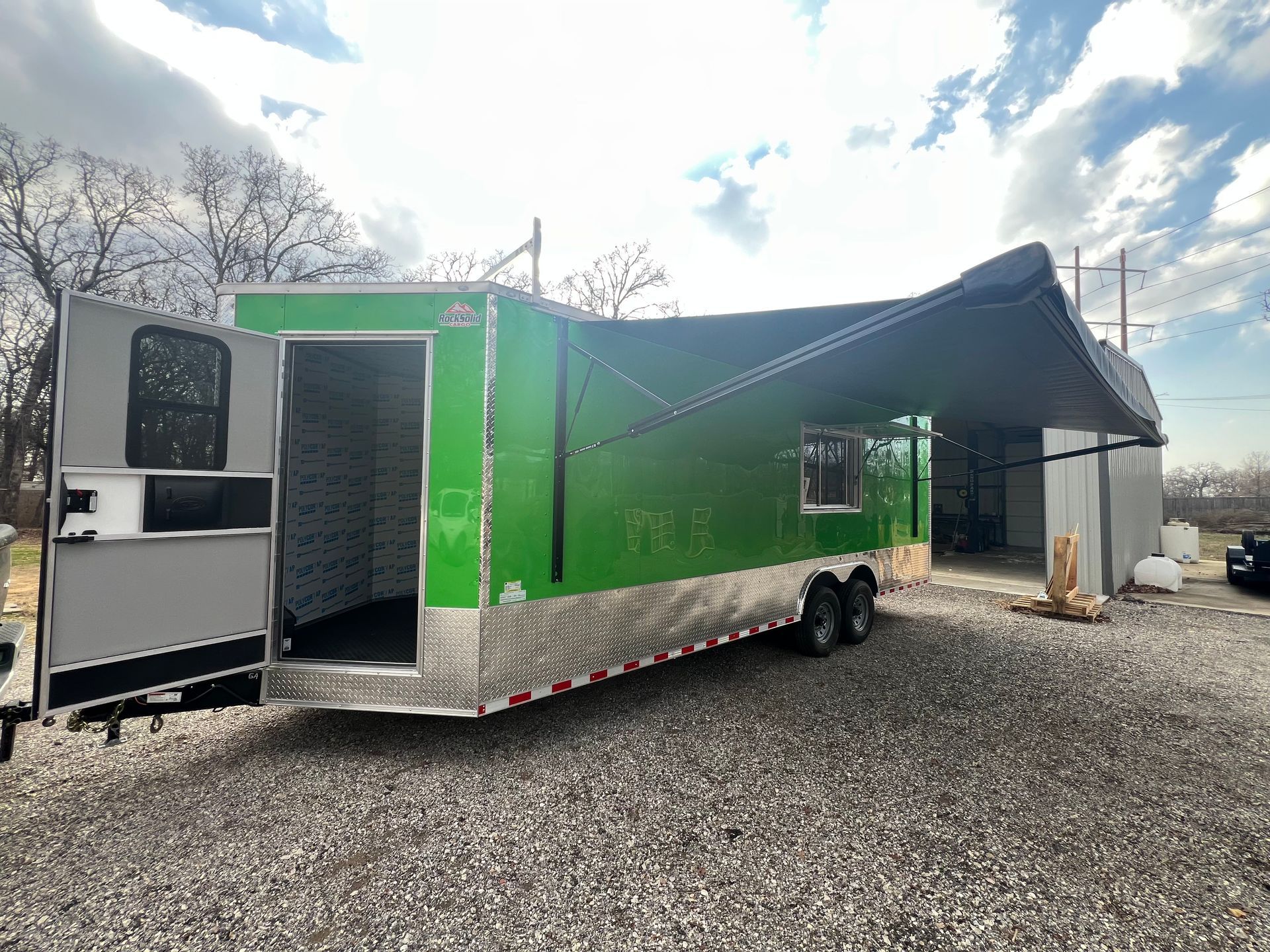 Green mobile food trailer with open side awning parked on gravel lot under cloudy sky