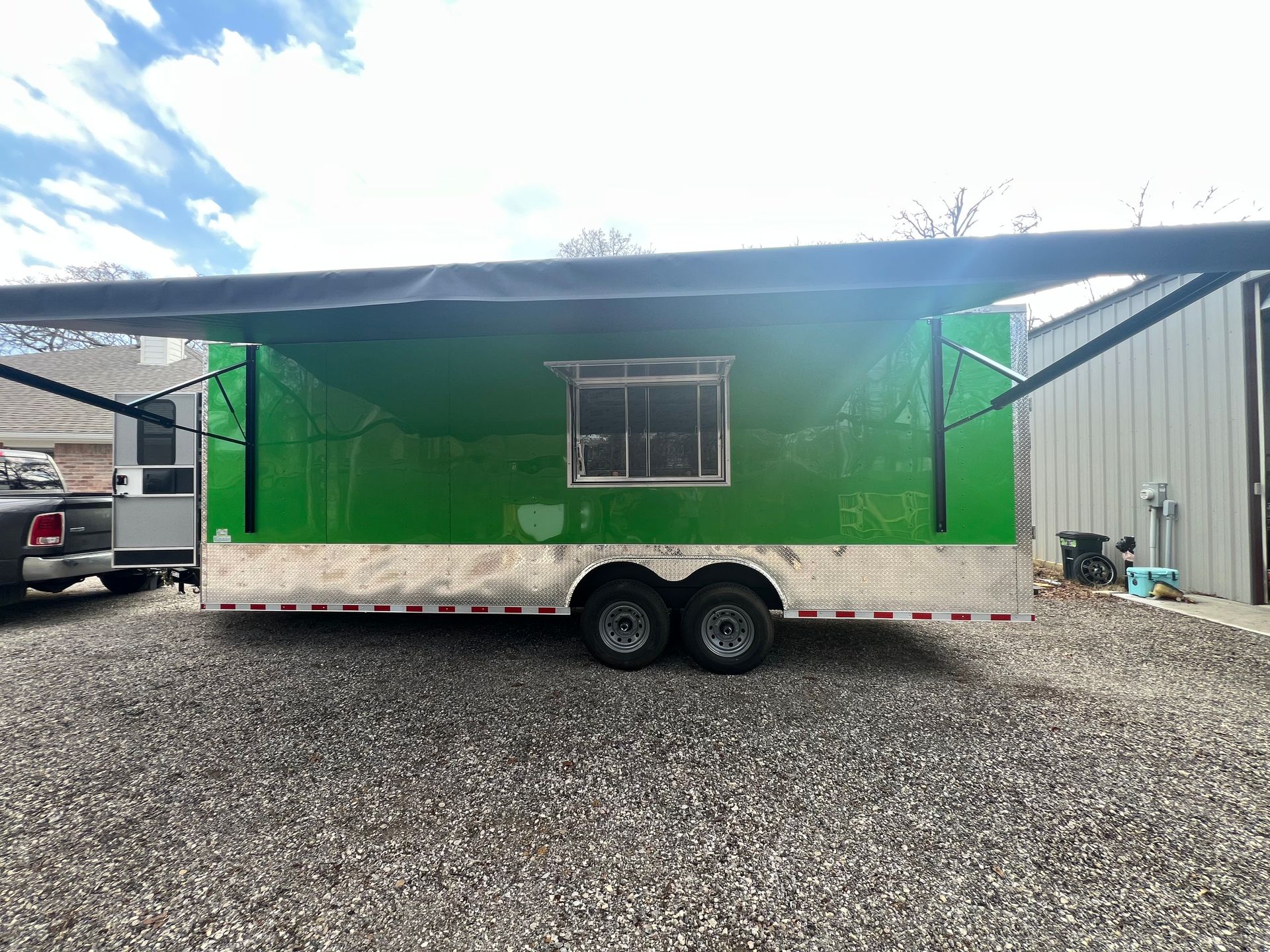 Green-and-white utility trailer with a window, parked on gravel beside industrial buildings