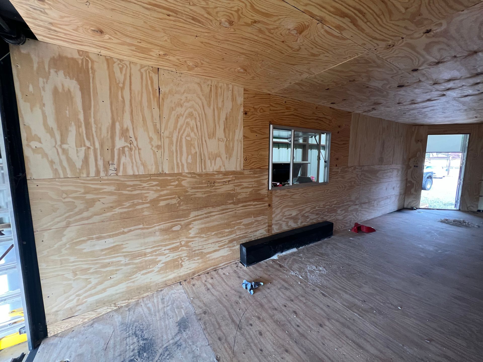 Plywood interior of a cargo trailer under upgrade construction with a window, doorway, and materials on the floor