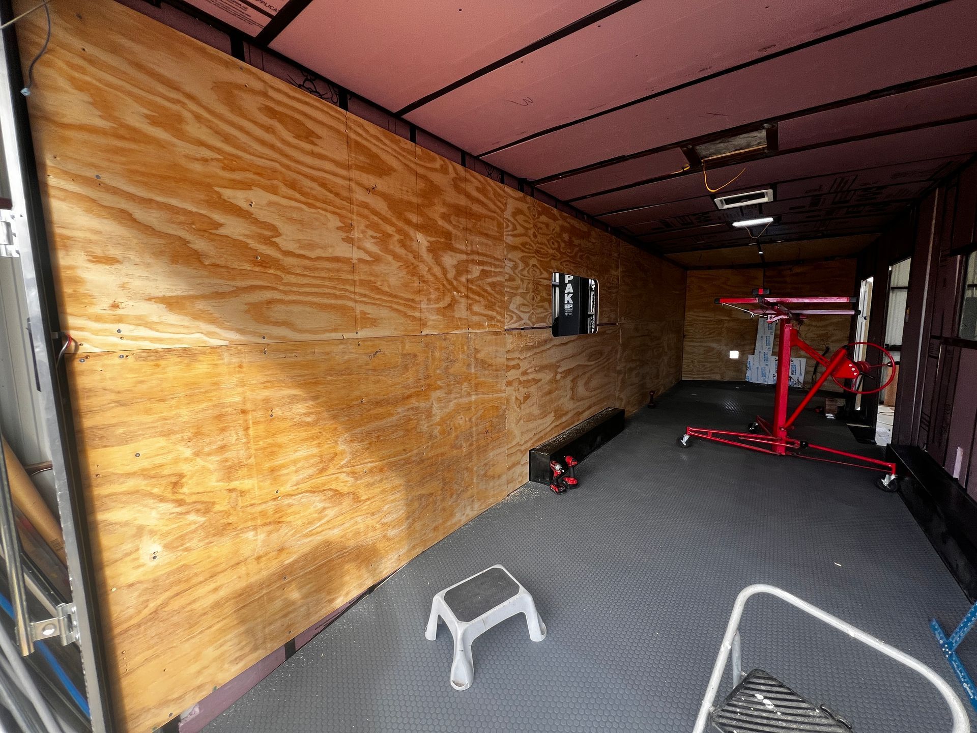 Empty cargo trailer interior with plywood walls, workout equipment, and a gray step stool on the floor