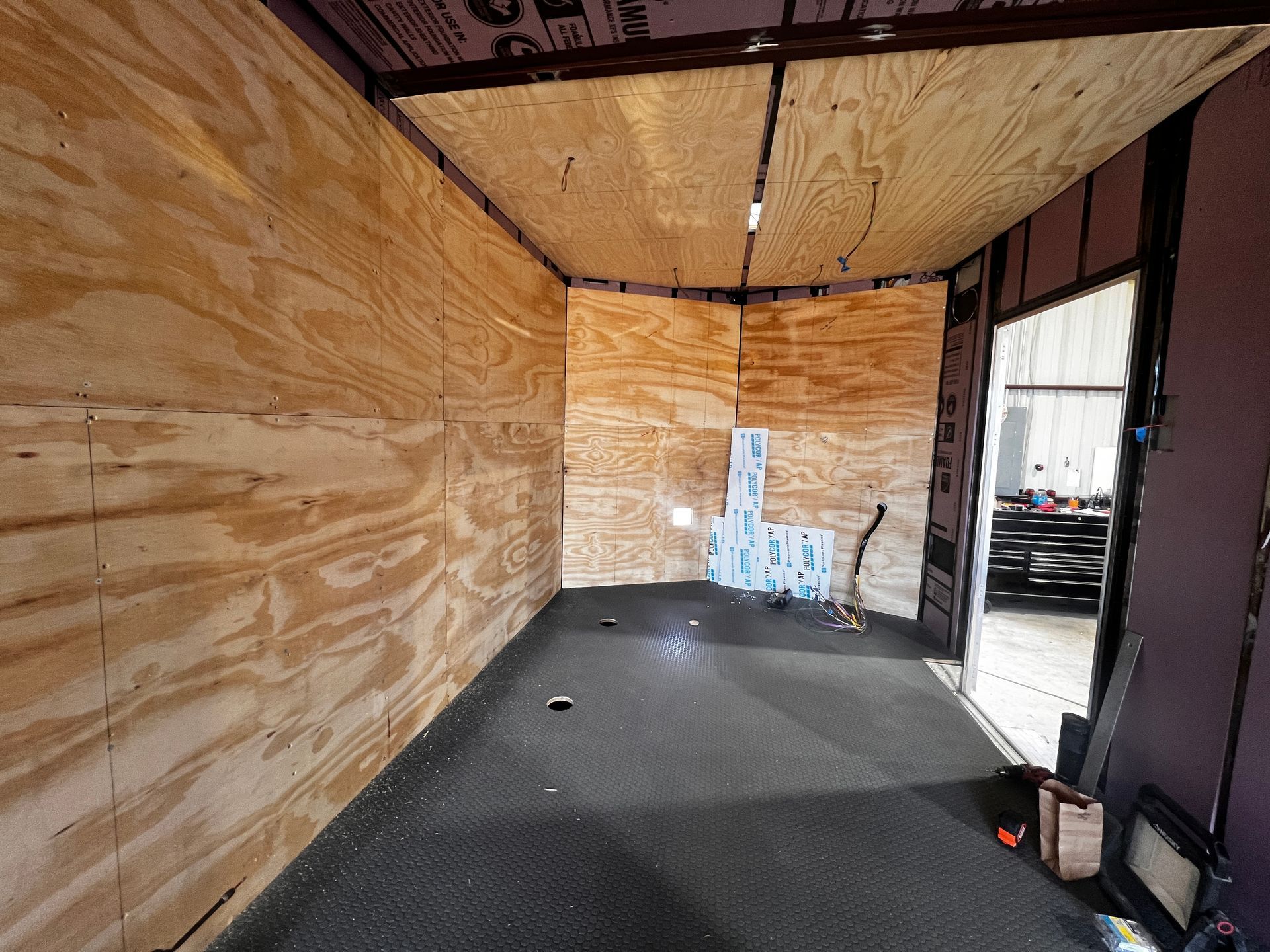 Empty plywood-lined truck or trailer interior with a black floor and open doorway on the right.