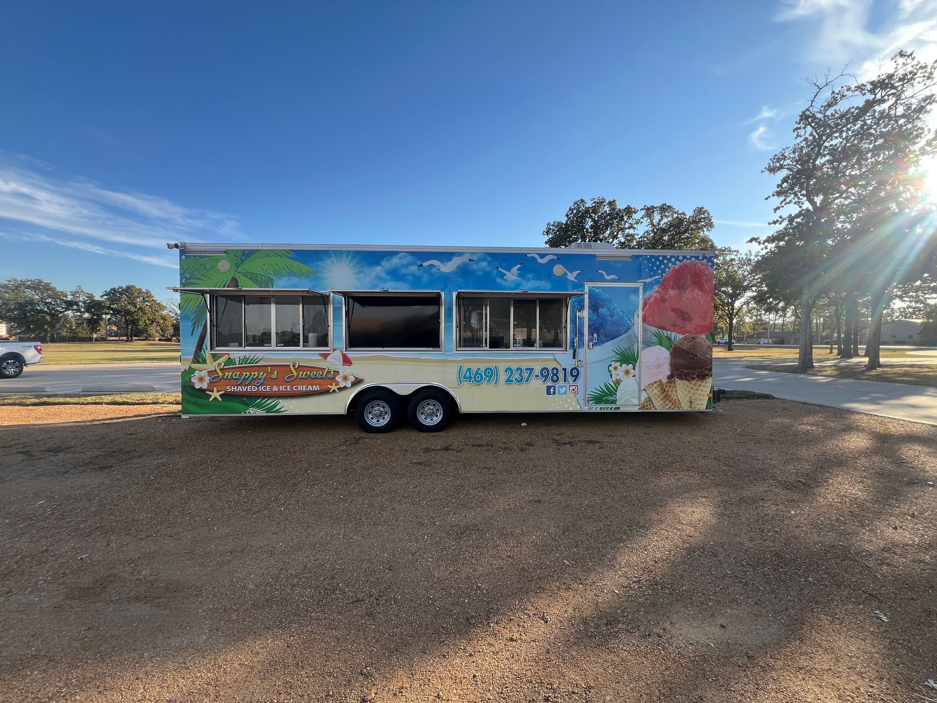 Colorful ice cream truck parked on a gravel lot under a blue sky, with serving windows open