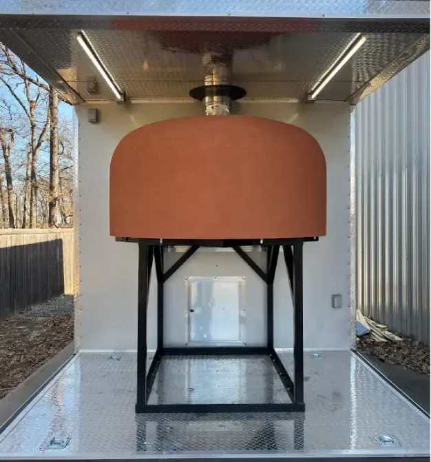 Large orange dome-shaped outdoor pizza oven on a black metal frame inside mobile food trailer.