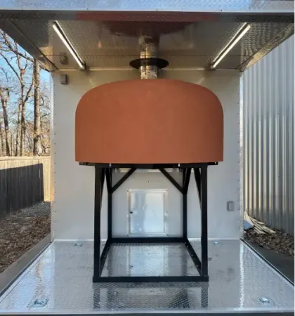 Large orange dome-shaped outdoor pizza oven on a black metal frame inside mobile food trailer.