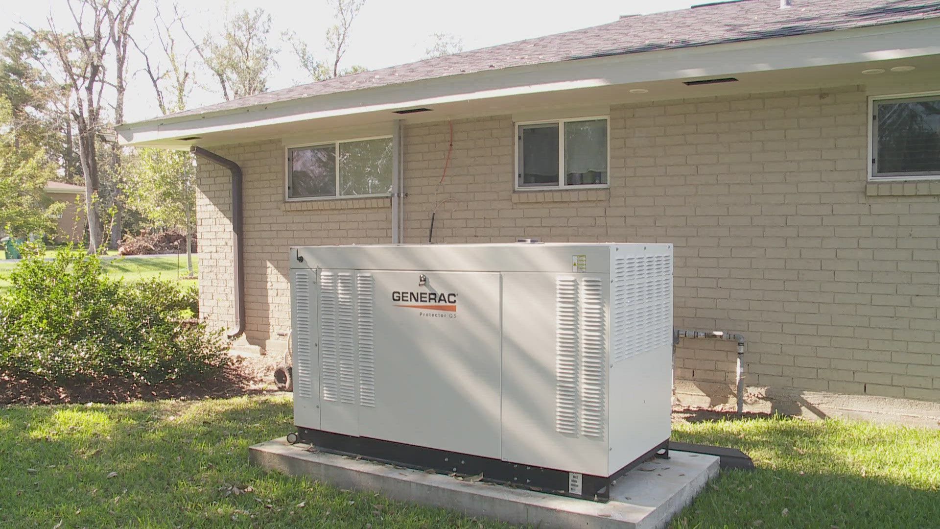Outdoor generator beside a beige house on a concrete pad, surrounded by grass and shrubs.