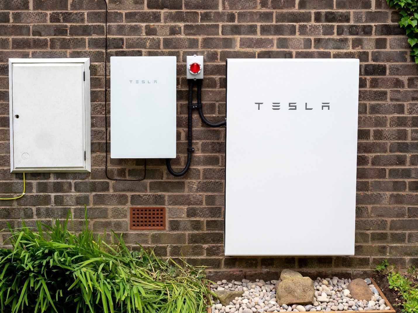 Tesla Powerwall and electrical boxes mounted on a brick exterior wall beside greenery
