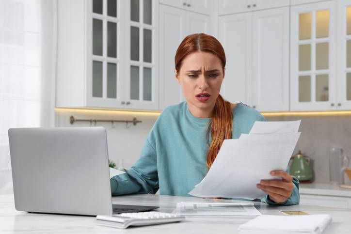 Woman at kitchen table reading bills with a laptop, looking worried