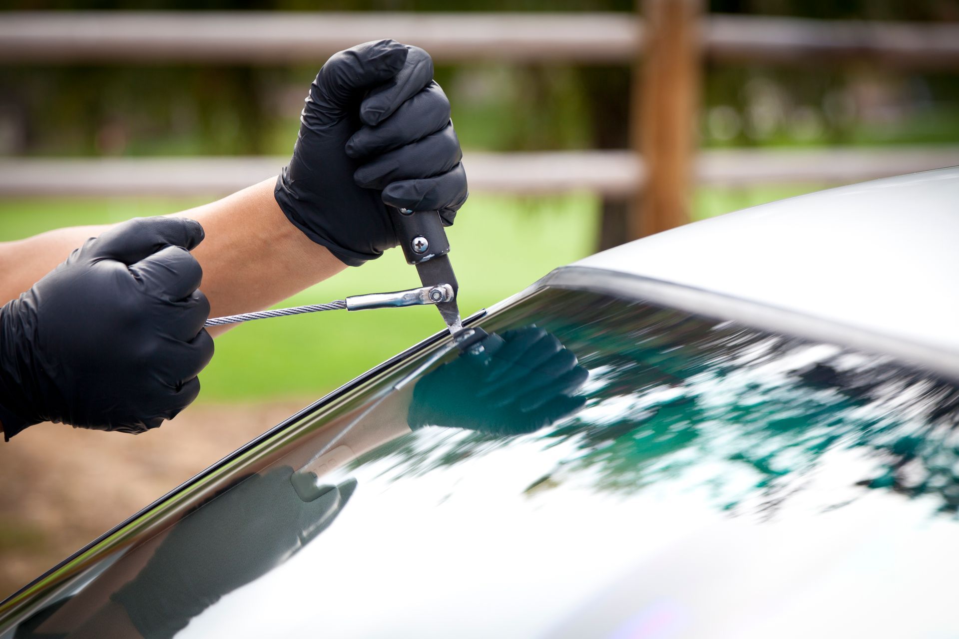 Hands in black gloves using tools to remove a car windshield in an outdoor setting.